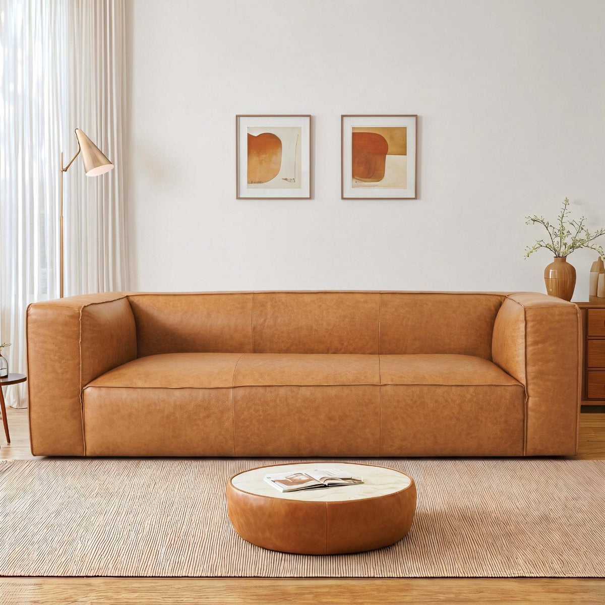 A modern tan leather sofa in a bright, minimalist living room with a round coffee table, wall art, and a neutral rug.