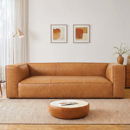 A modern tan leather sofa in a bright, minimalist living room with a round coffee table, wall art, and a neutral rug.