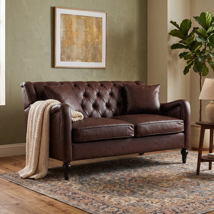 A Dark Brown Loveseat leather loveseat with tufted button detailing on the backrest and seat, styled in a living room setting with a cream knit throw blanket, a patterned area rug, a wooden side table, and a potted fiddle-leaf fig plant.