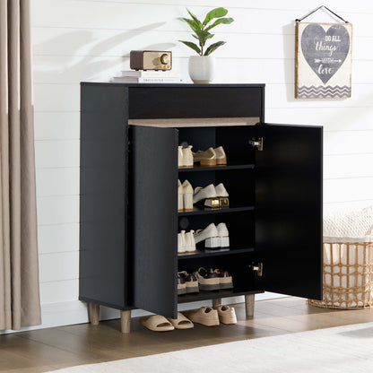A black shoe cabinet with open doors, shown in a styled room setting with shoes stored on shelves and a few placed on the floor. The cabinet has a top drawer, a plant on top, and is set against a white shiplap wall.