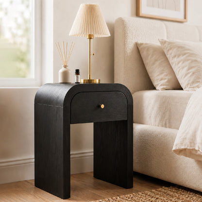 A modern black wooden nightstand with a single curved drawer and a small round brass knob, styled next to a bed in a bright bedroom. A table lamp and a reed diffuser sit on top of the nightstand.