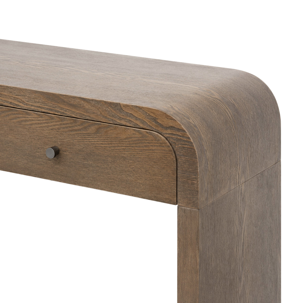Close-up of a wooden table corner showing a medium brown wood grain finish, rounded apron, and a small round dark metal drawer knob on a white background.