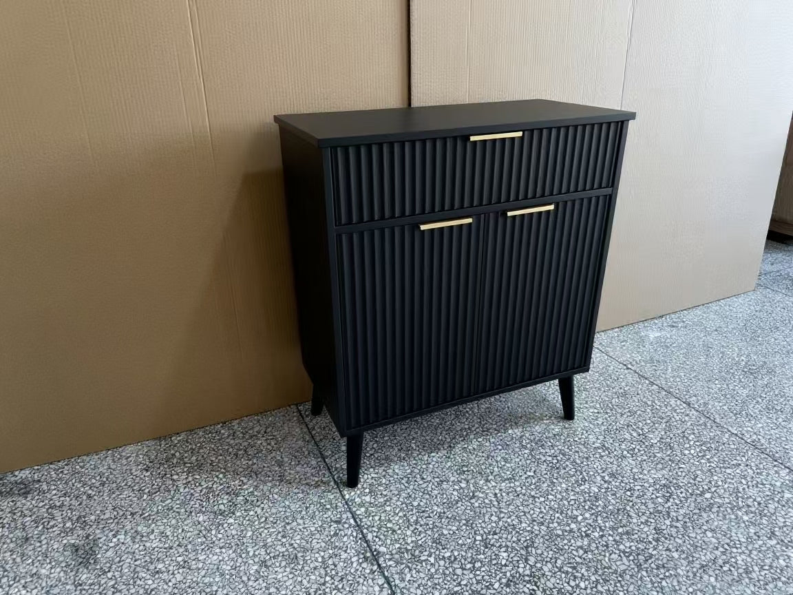 A black ribbed cabinet with two drawers and two doors, featuring slim gold metal handles and tapered black legs, positioned against a cardboard box backdrop on a speckled gray floor.