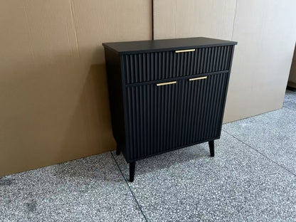 A black ribbed cabinet with two drawers and two doors, featuring slim gold metal handles and tapered black legs, positioned against a cardboard box backdrop on a speckled gray floor.