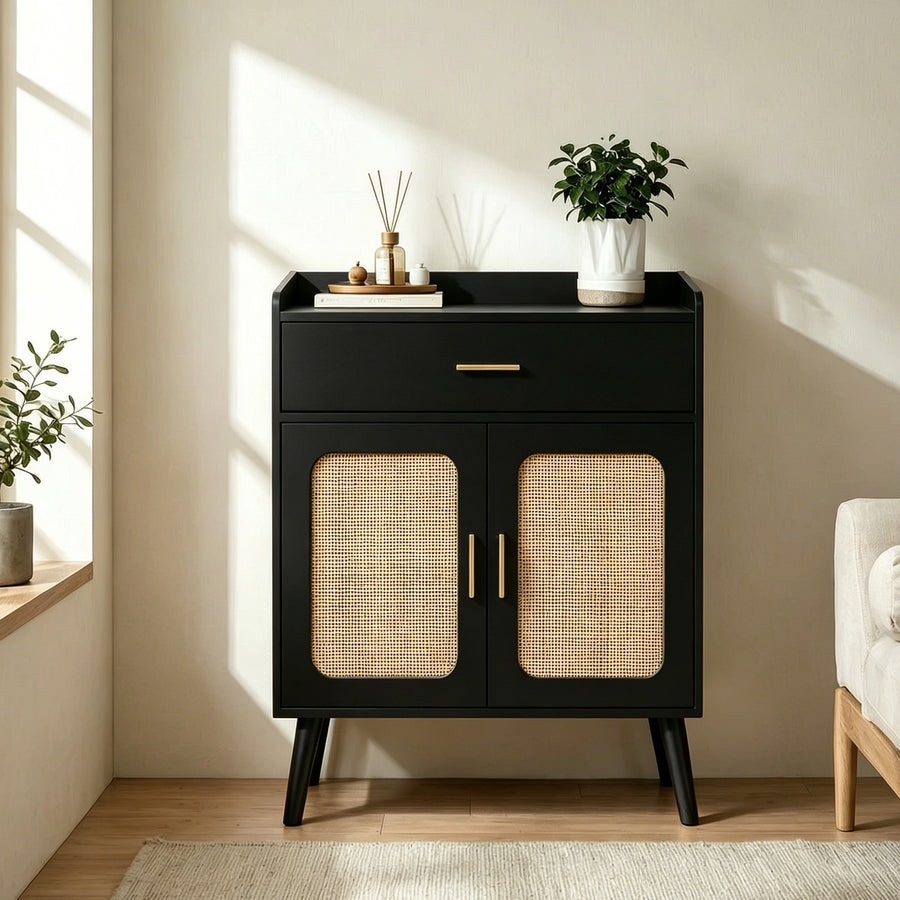 A modern black side cabinet with two doors featuring woven cane panels and gold-tone handles, styled in a sunlit room with a plant and diffuser on top.
