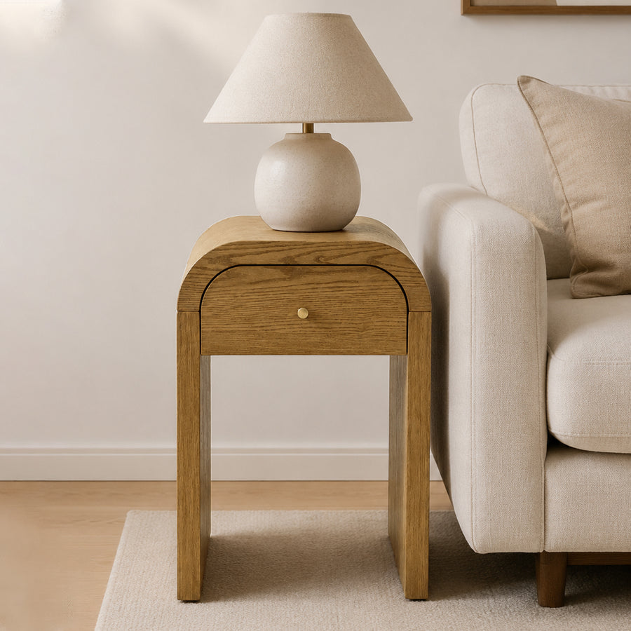 A light Walnut Brown oak wood end table with a single curved drawer and a small brass knob, styled next to a cream-colored sofa in a bright, modern living room. A matching ceramic table lamp sits on top of the table.