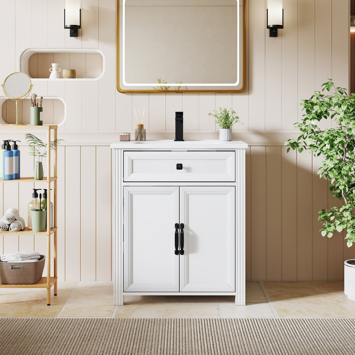 A white bathroom vanity cabinet with two doors and black handles, paired with a white sink and black faucet, set against a beige paneled wall with a gold-framed mirror, wall sconces, a shelving unit, and a potted plant.