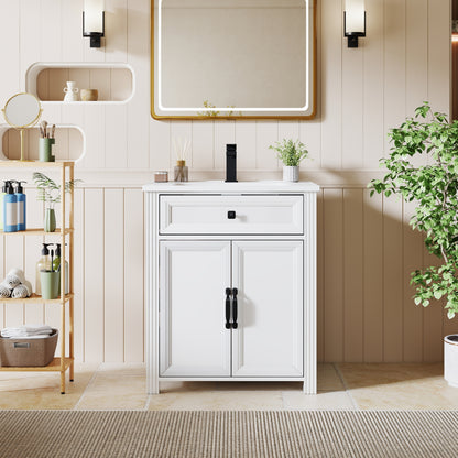 A white bathroom vanity cabinet with two doors and black handles, paired with a white sink and black faucet, set against a beige paneled wall with a gold-framed mirror, wall sconces, a shelving unit, and a potted plant.