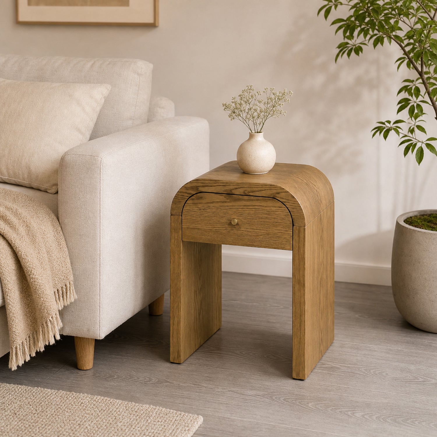 A light Walnut Brown oak wood end table with a single drawer and rounded top, styled in a cozy living room next to a cream armchair with a small white vase on top.