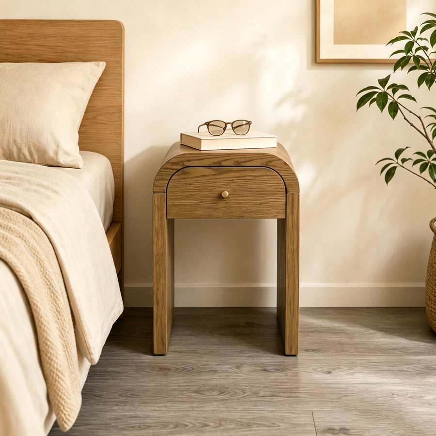 A light oak wood Walnut Brown nightstand with a single curved drawer and a small brass knob, positioned next to a bed in a warm, neutral-toned bedroom. A book and a pair of glasses sit on top of the nightstand, and a potted plant is visible on the right.