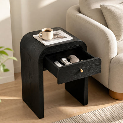 A black wooden nightstand with a curved top and an open drawer, styled in a living room next to a beige armchair with a coffee mug on top.