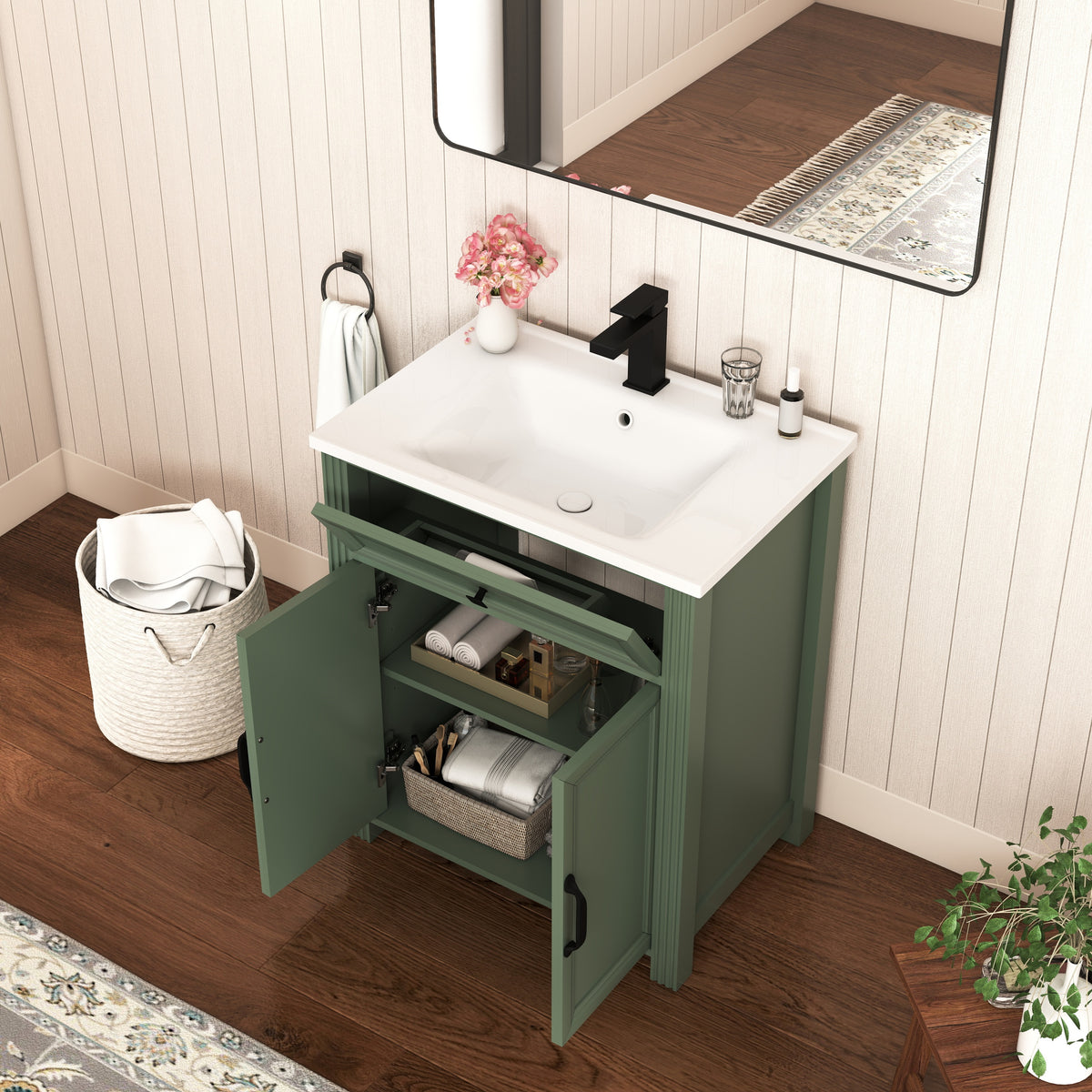 Olive green bathroom vanity cabinet with a sink countertop and black faucet, shown in a styled bathroom setting with an open door revealing storage shelves, a mirror, and wood flooring.