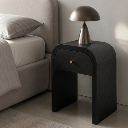 A modern black nightstand with a single drawer and a small gold knob, styled next to a bed in a minimalist bedroom. A dome-shaped table lamp with a brushed silver base sits on top of the nightstand.
