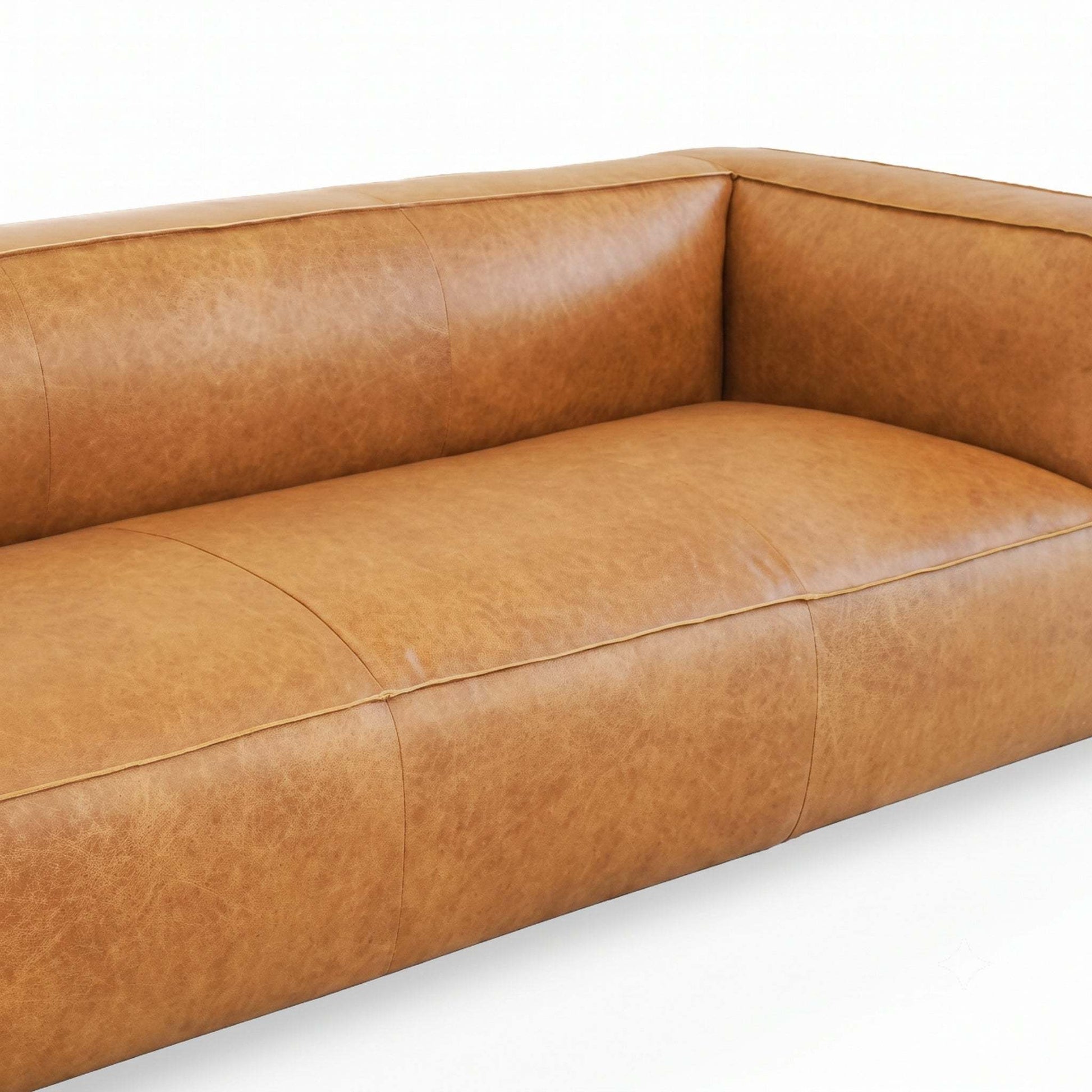A close-up of a modern leather sofa in a warm tan or camel brown color, showing the textured leather upholstery, channel tufting on the backrest, and clean stitching on a white background.