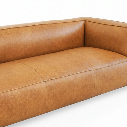 A close-up of a modern leather sofa in a warm tan or camel brown color, showing the textured leather upholstery, channel tufting on the backrest, and clean stitching on a white background.
