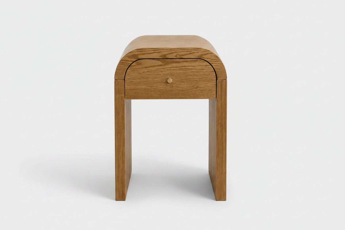 A modern Walnut Brown wooden nightstand with a curved top, a single drawer featuring a small round brass knob, and four tapered legs, shown against a light gray background.
