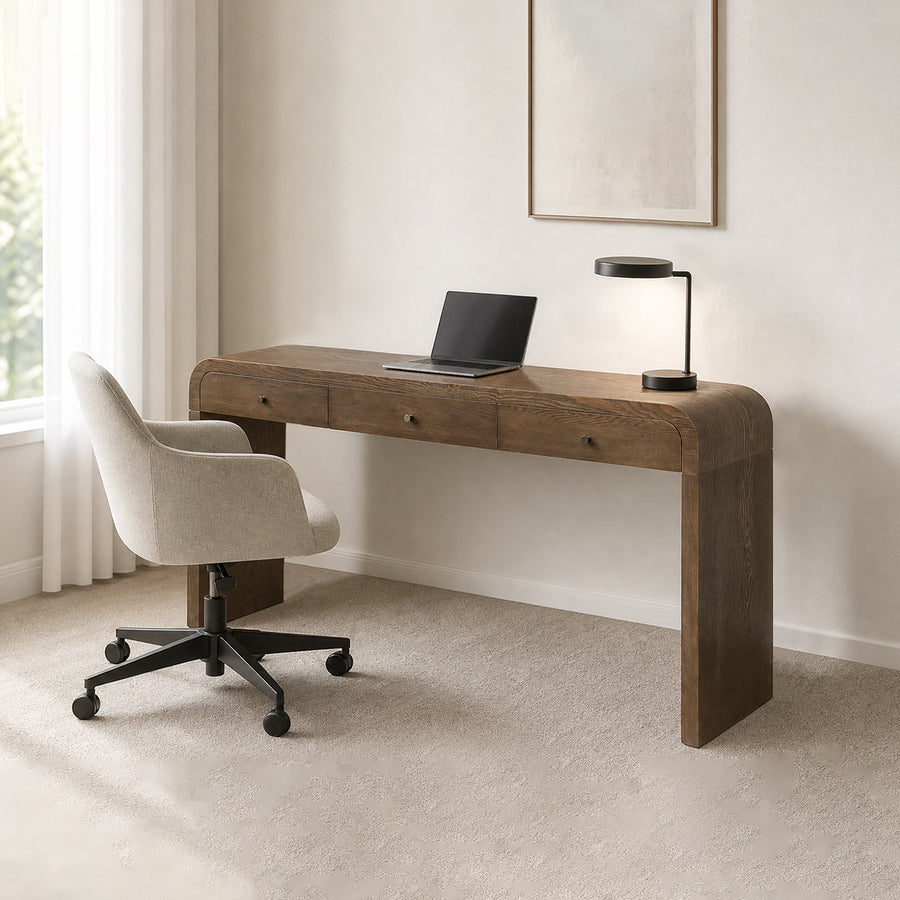 A modern writing desk in a warm medium brown wood finish, styled in a minimalist home office with a beige upholstered rolling office chair, a black desk lamp, and a closed laptop on top.