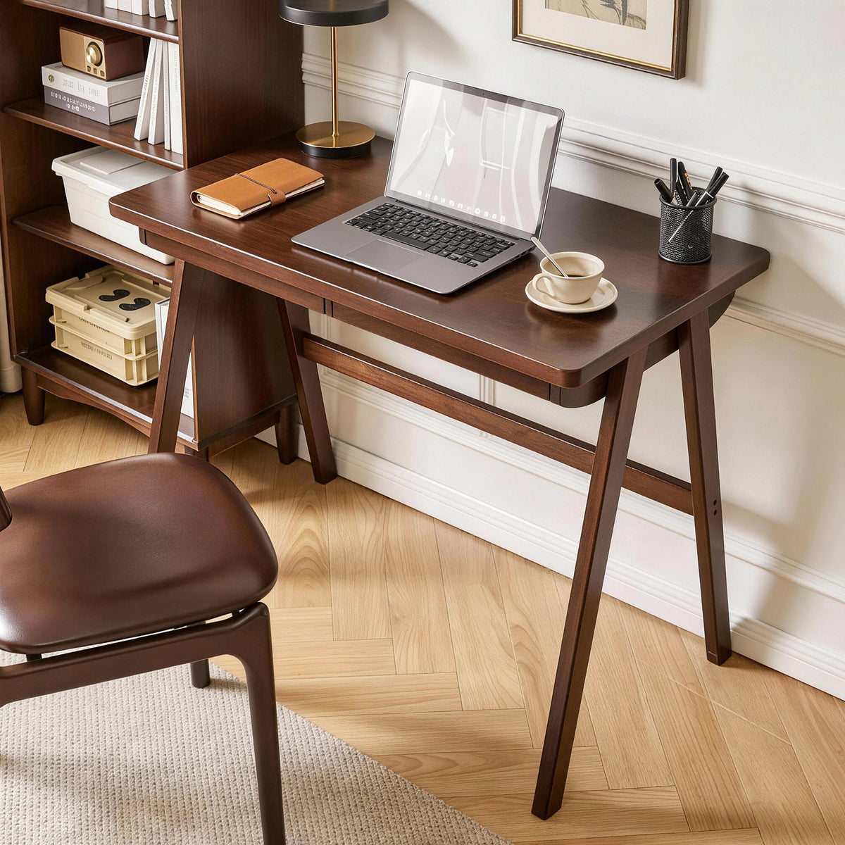 A dark brown wooden writing desk with angled legs and a matching wooden chair in a home office setting, styled with a laptop, notebook, lamp, and coffee cup.