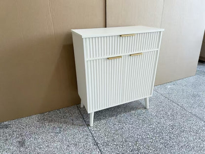 A white mid-century modern cabinet with fluted vertical panels, two drawers on top and two doors below, accented with gold-tone handles and legs, set against a cardboard box backdrop on a speckled gray floor.