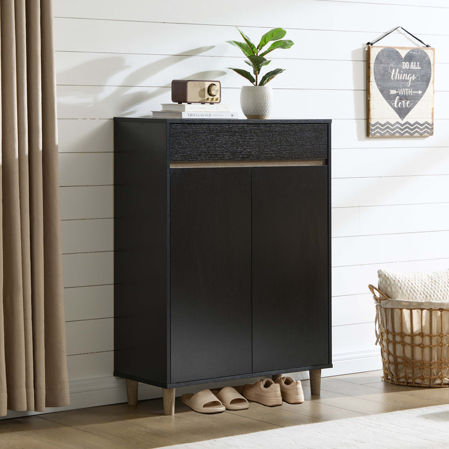 A tall black entryway cabinet with two doors and a textured top drawer section, styled in a bright room with a plant, books, a small radio, slippers, a curtain, and a basket nearby.