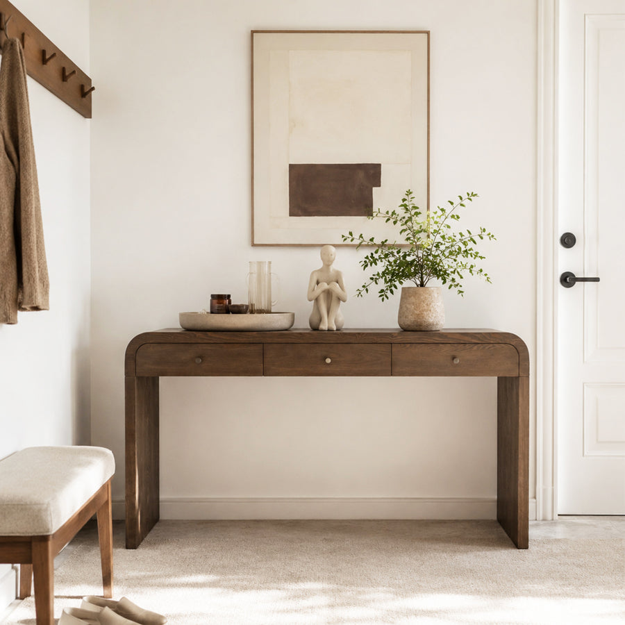 A dark brown wooden console table styled in a bright, modern entryway with a potted plant, ceramic figurine, and glass vases, set against a white wall with a minimalist framed print.