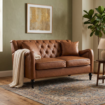 A brown leather loveseat sofa with tufted button detailing, shown in a styled living room setting with a patterned area rug, a cream knit throw blanket, a side table, and a potted plant. light brown loveseat