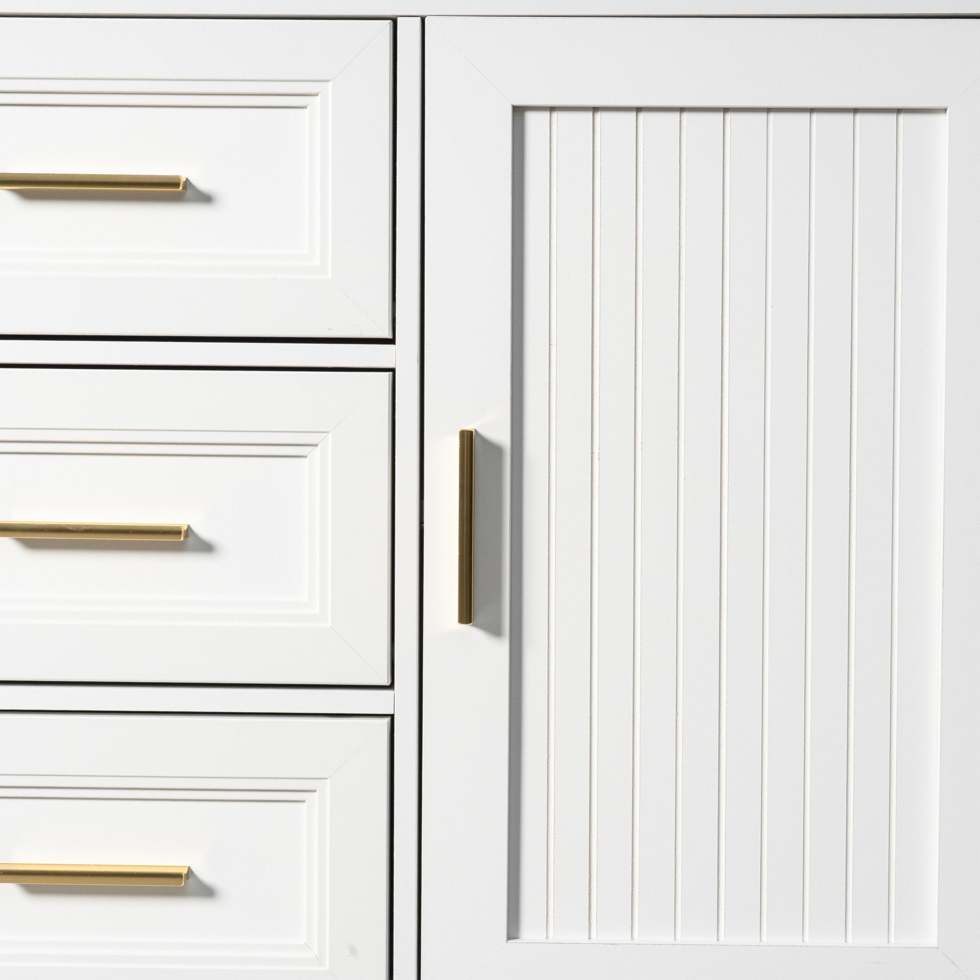 Close-up of a white cabinet with a vertical beadboard door panel and a slim vertical gold handle, next to white drawers with horizontal gold handles.
