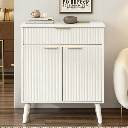 A modern white sideboard cabinet with vertical fluted panel doors and drawers, featuring slim gold metal handles and tapered white legs, styled in a contemporary living room setting.