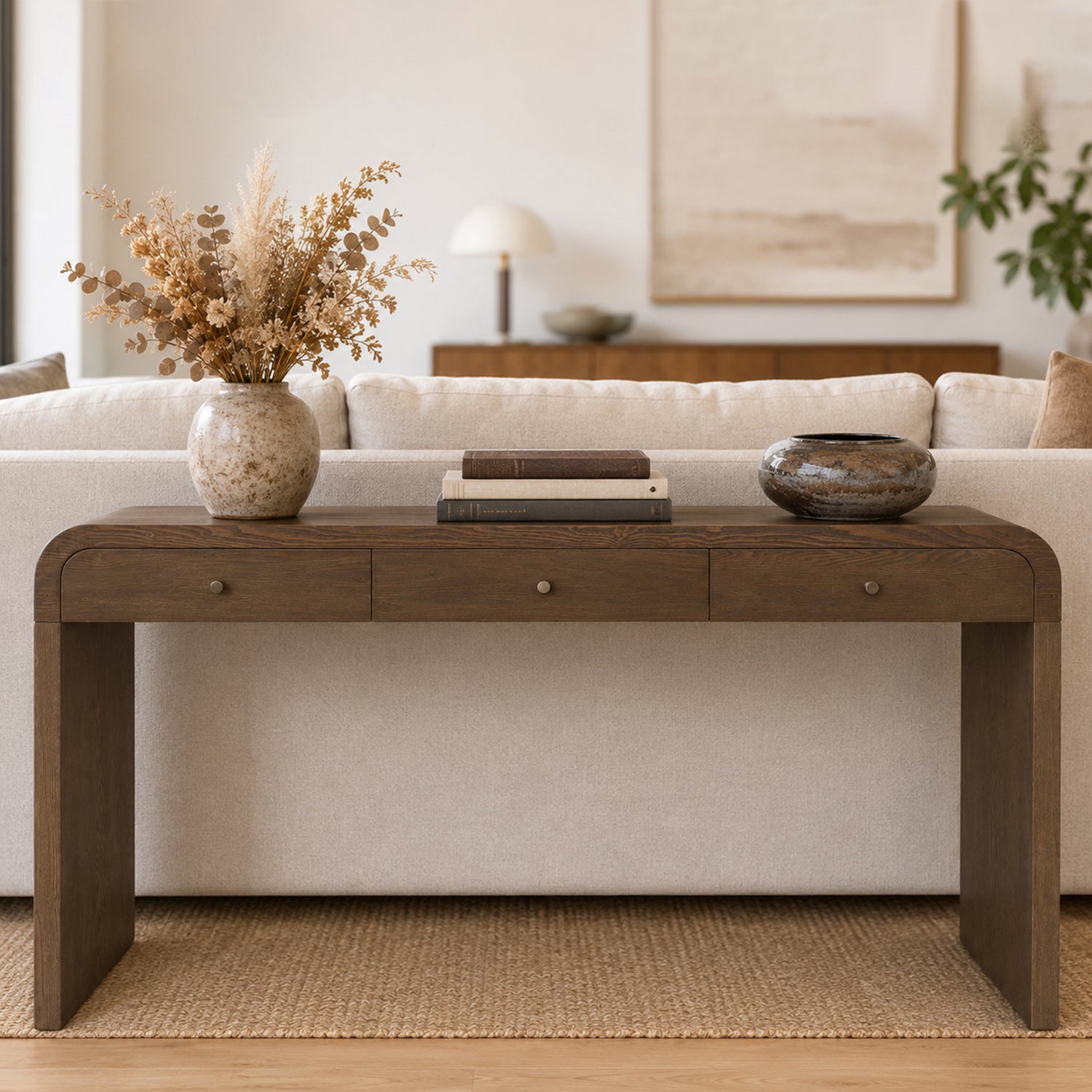 A dark brown wooden console table in a modern living room setting, styled with a vase of dried grass, books, and a decorative bowl on top.
